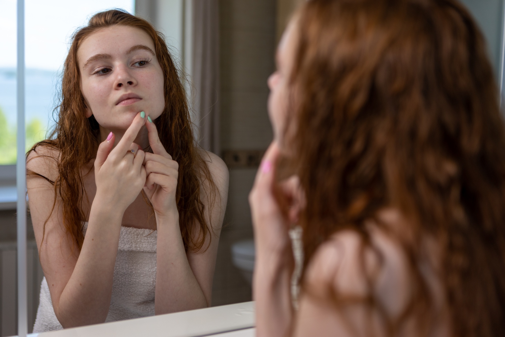 A young girl squeezes pimples while standing in the bathroom at the mirror